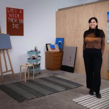 Mia Boe standing on striped rugs on a concrete floor, in a room with scattered paintings around the walls