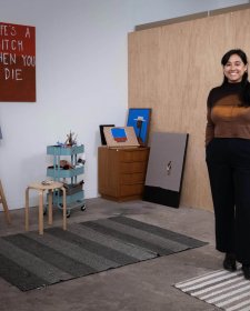 Mia Boe standing on striped rugs on a concrete floor, in a room with scattered paintings around the walls