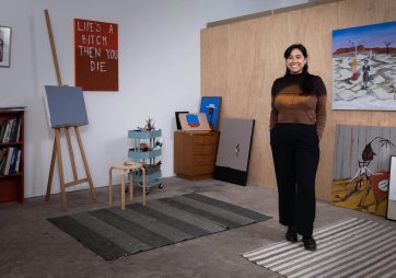 Mia Boe standing on striped rugs on a concrete floor, in a room with scattered paintings around the walls