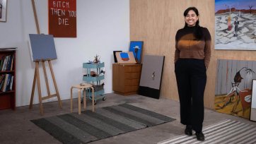 Mia Boe standing on striped rugs on a concrete floor, in a room with scattered paintings around the walls