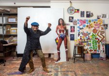 Kaylene Whiskey in her studio, raising her arms in the air in celebration, next to a life size cutout of wonder woman and an artwork in progress