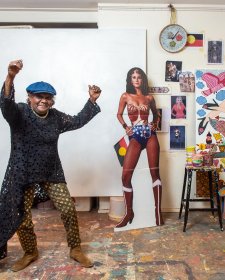 Kaylene Whiskey in her studio, raising her arms in the air in celebration, next to a life size cutout of wonder woman and an artwork in progress