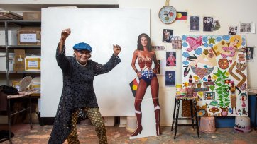 Kaylene Whiskey in her studio, raising her arms in the air in celebration, next to a life size cutout of wonder woman and an artwork in progress