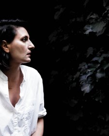 Angelica Mesiti in profile wearing a white linen shirt in front of a dark leafy hedge