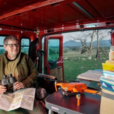 Julie Gough sitting in the back of a red vehicle while holding binoculars, with sheep grazing in a field in the background