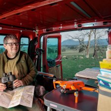 Julie Gough sitting in the back of a red vehicle while holding binoculars, with sheep grazing in a field in the background