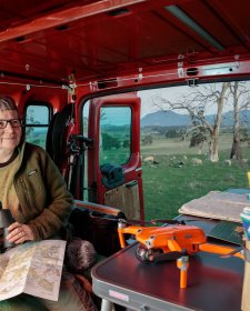 Julie Gough sitting in the back of a red vehicle while holding binoculars, with sheep grazing in a field in the background