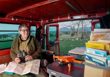 Julie Gough sitting in the back of a red vehicle while holding binoculars, with sheep grazing in a field in the background