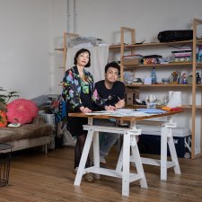 Two people at a bench covered in sheets of paper in a room with a wooden floor and a large brown couch