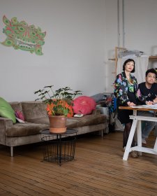 Two people at a bench covered in sheets of paper in a room with a wooden floor and a large brown couch