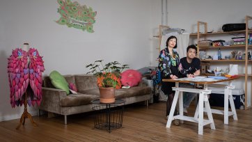 Two people at a bench covered in sheets of paper in a room with a wooden floor and a large brown couch