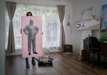 Baby Guerrilla standing behind a large artwork in a room with a wooden floor and a large window looking onto a leafy green garden