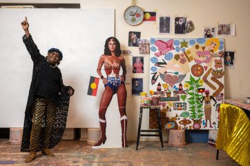 Kaylene Whiskey in her studio, raising her arms in the air in celebration, next to a life size cutout of wonder woman and an artwork in progress