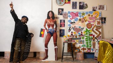 Kaylene Whiskey in her studio, raising her arms in the air in celebration, next to a life size cutout of wonder woman and an artwork in progress Kaylene Whiskey in her studio, raising her arms in the air in celebration, next to a life size cutout of wonder woman and an artwork in progress