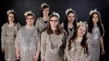 7 smiling, dark haired young women standing in front of a black background. They are wearing small cream and gold head dresses and pale gold sequinned dresses which sparkle in the light.