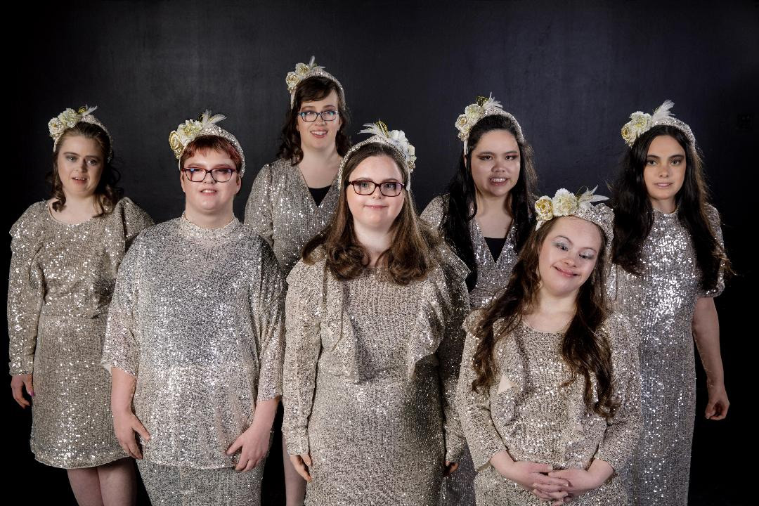 7 smiling, dark haired young women standing in front of a black background. They are wearing small cream and gold head dresses and pale gold sequinned dresses which sparkle in the light.