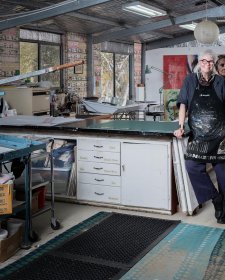 Alison Alder wearing a paint splattered apron, leaning against a bench in her studio