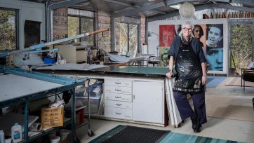 Alison Alder wearing a paint splattered apron, leaning against a bench in her studio