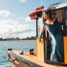 A person with long hair being blown by the wind, wearing a blue dress, while leaning over the edge of a boat on the water