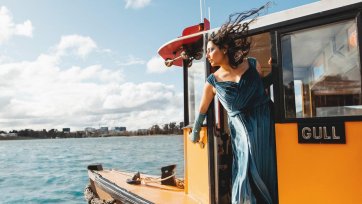 A person with long hair being blown by the wind, wearing a blue dress, while leaning over the edge of a boat on the water