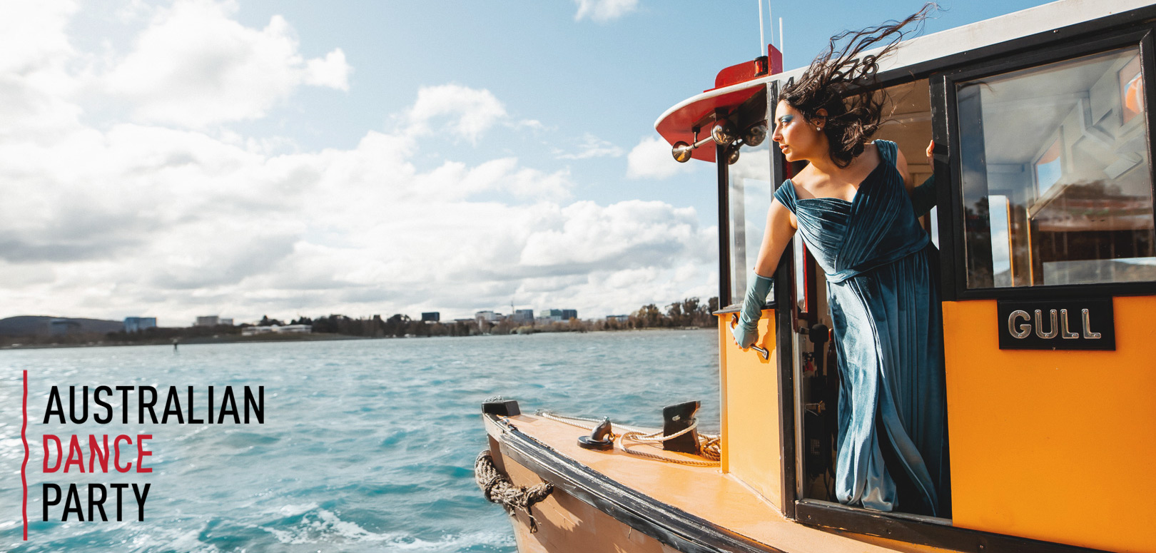 A person with long hair being blown by the wind, wearing a blue dress, while leaning over the edge of a boat on the water
