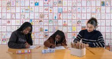 Three people drawing at a wooden table in front of a wall of drawings