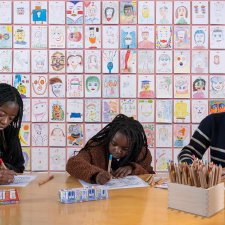 Three people drawing at a wooden table in front of a wall of drawings