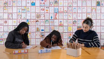 Three people drawing at a wooden table in front of a wall of drawings